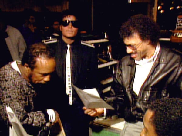 Michael Jackson, wearing a black shirt, white tie and sunglasses looks ahead as Quincy Jones leans in on a piano and Lionel Richie smiles while holding a piece of paper.