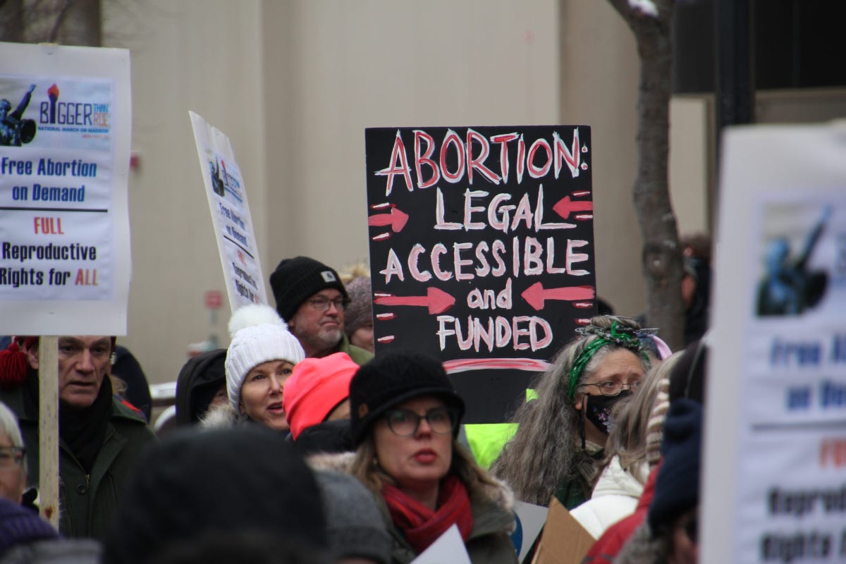 A photo from a protest, showing a crowd of people wearing cold-weather clothing and holding signs. The most visible says, “Abortion: Legal, Accessible, and Funded” in blended red and white letters on a black backdrop.