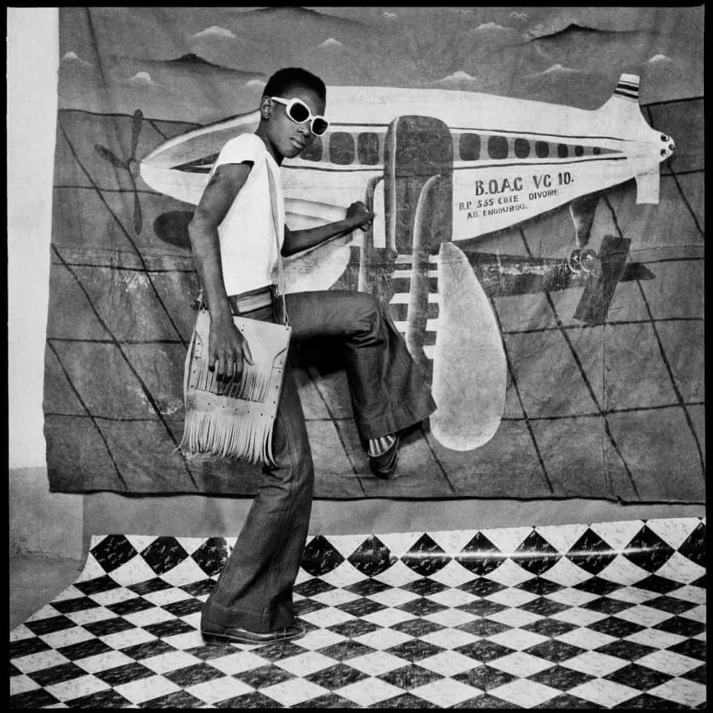 A painted backdrop of a plane in a photo studio, a young man pretends to climb into this backdrop wearing sunglasses and a handbag