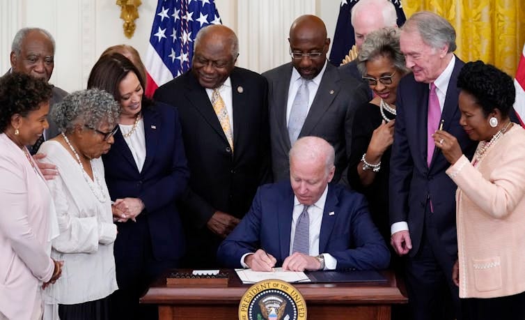 A an at a desk signing a piece of paper with many people standing around him, watching. A an at a desk signing a piece of paper with many people standing around him, watching.