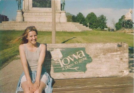 young Karrie with long dirty blonde hair blowing in the wind, seated on a bench painted with the old Iowa state logo "A Place to Grow" and smiling.