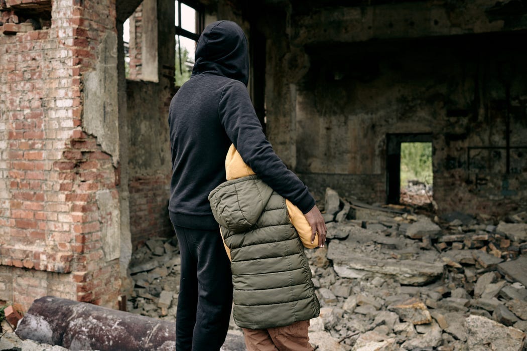 Man and child standing before rubble, what used to be a building. Their backs are to the camera — the man’s arm resting protectively on the child’s shoulder.