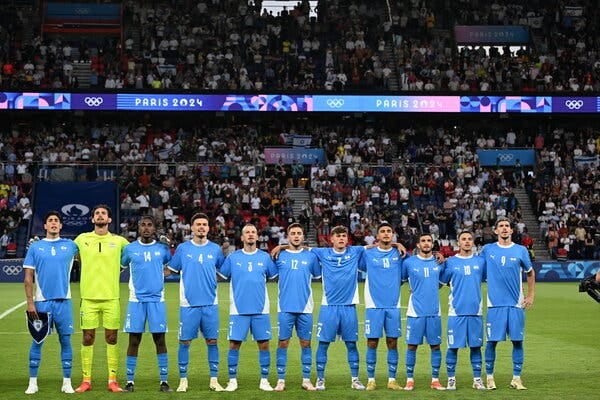 Members of Israel’s soccer team, dressed in light blue jerseys except for a goalkeeper in green, stand arm in arm and sing their national anthem before a match.