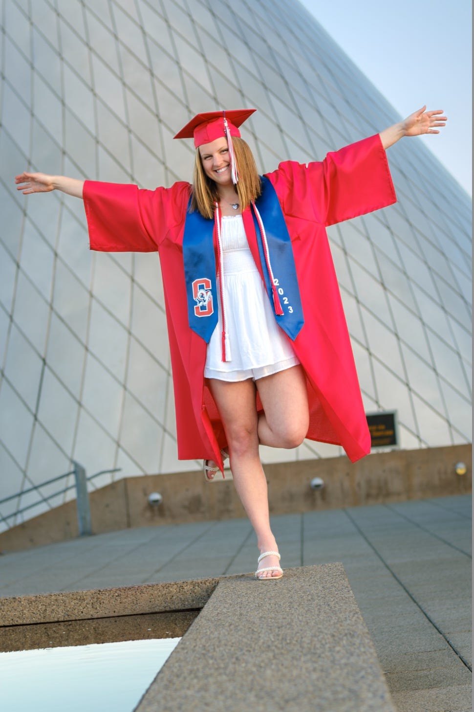A young woman wearing a red cap and gown and a blue sash balances on one leg in front of the metal cone sculpture at the Museum of Glass.
