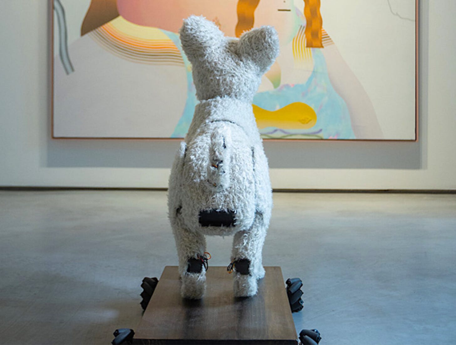 A view from behind a toy dog. The dog is on a platform on wheels and it's considering an artwork in front of it 
