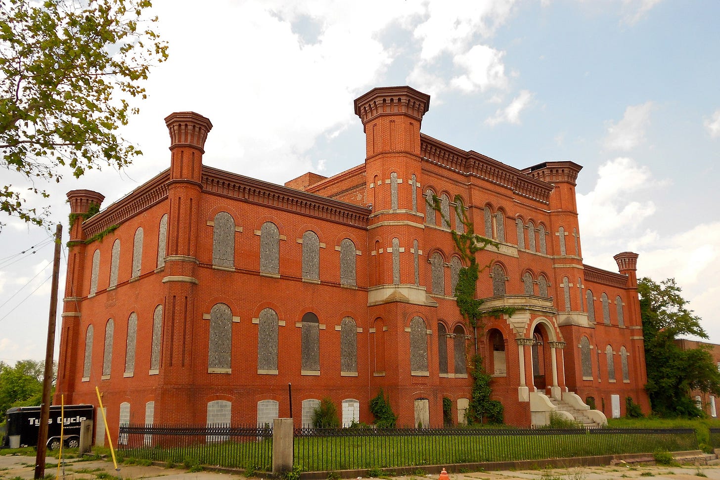 The front side of a large brick building with boarded up windows viewed from the street. It is accented on its corners with ornate brick pillars.