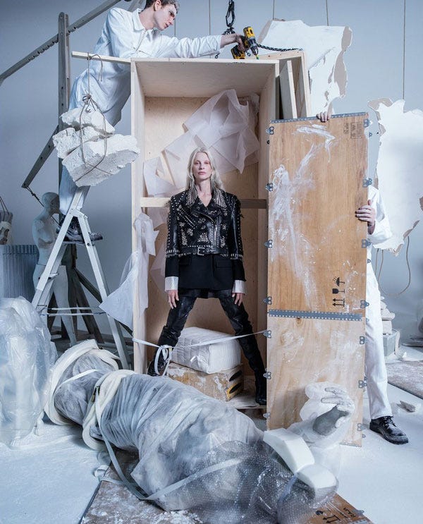 A fashion ad. A blonde white woman in a black military style jacket stands in a wooden crate as two men unbox her. On the floor, a statue lies wrapped in plastic. Chunks of plaster fall from the ceiling and off the walls.