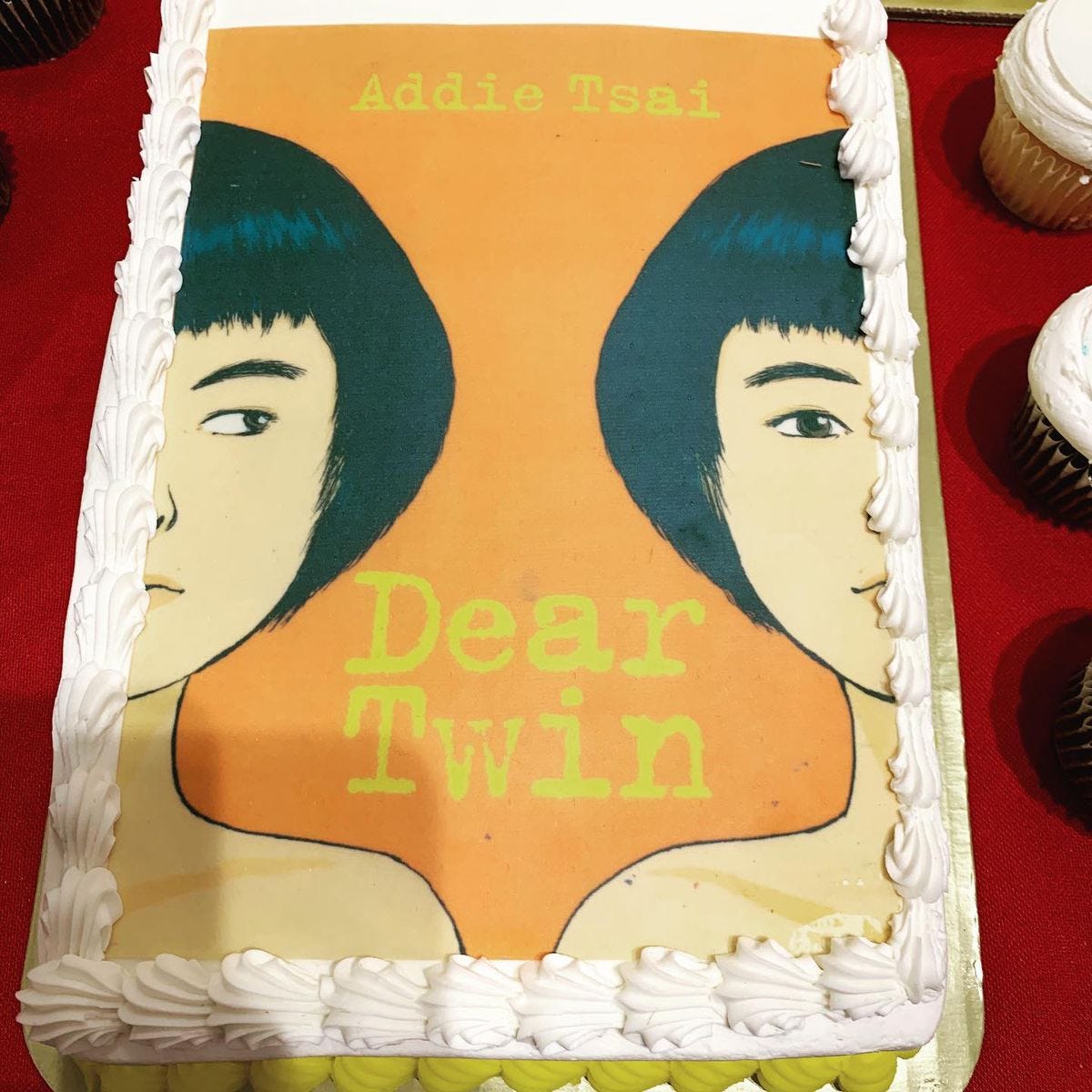 A cake decorated with the cover of Addie Tsai’s book, Dear Twin. It shows two people with short black hair and bangs side-by-side. Only half of each person is visible. The background is orange, with Addie Tsai’s name and Dear Twin in yellow text in a typewriter-style font.