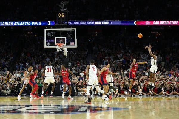 San Diego State’s Lamont Butler, in white at right, jumps in the air with his arms up and the ball flying toward the basket, with the other players on the court watching.