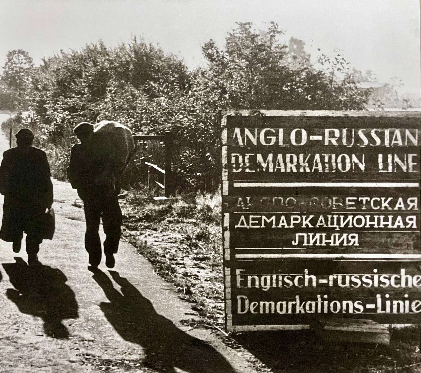 Two figures walk along a country path in silhouette, one carrying a bundle. A multilingual wooden sign reads “Anglo-Russian Demarkation Line” in English, Russian, and German, marking the post-war division of occupied Germany. Two figures walk along a country path in silhouette, one carrying a bundle. A multilingual wooden sign reads “Anglo-Russian Demarkation Line” in English, Russian, and German, marking the post-war division of occupied Germany.