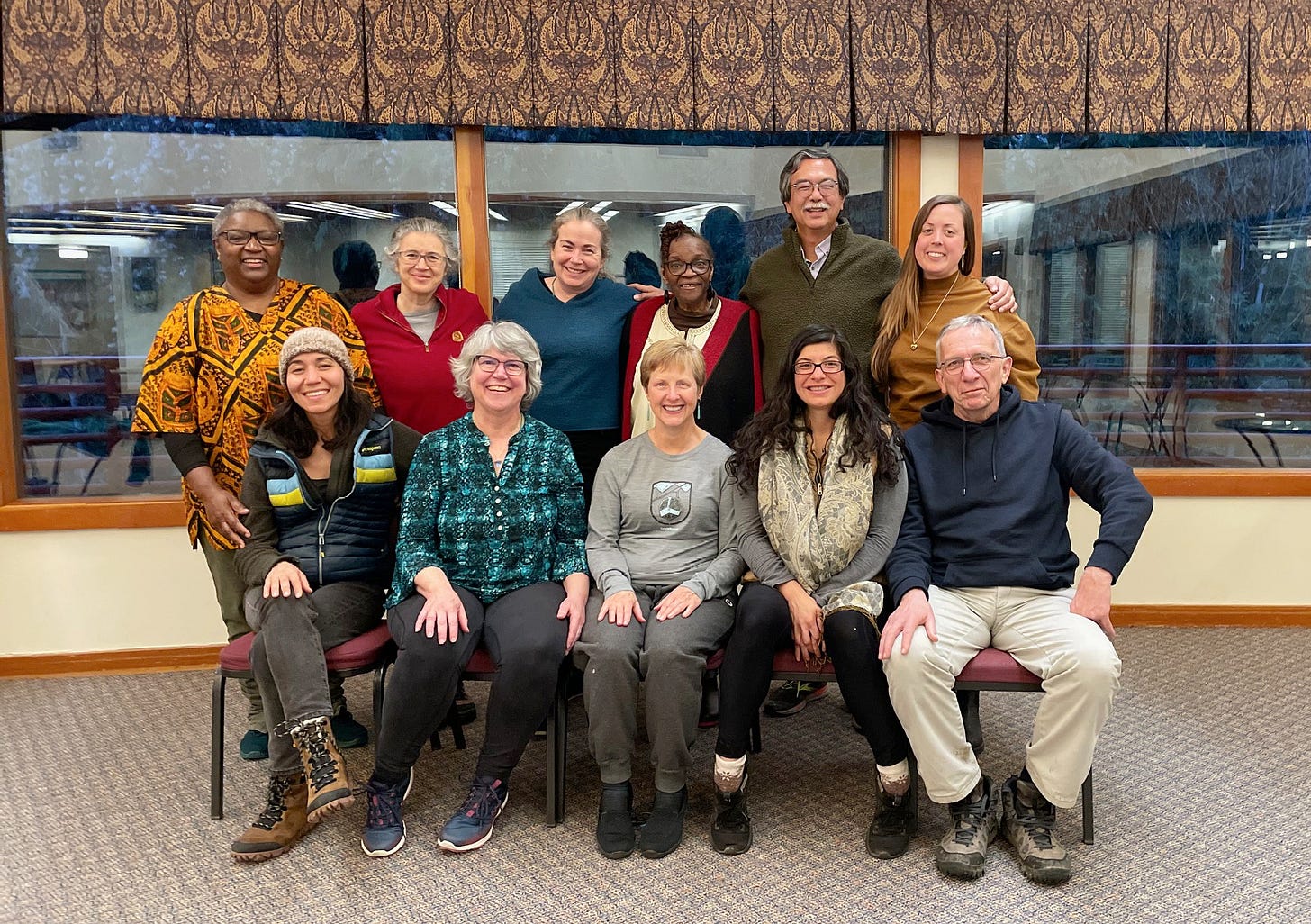 11 people posed in two rows, looking at camera. Smiling. Many different skin colors and different color clothes. In a room with carpet. Front row is sitting on chairs, back row is standing.