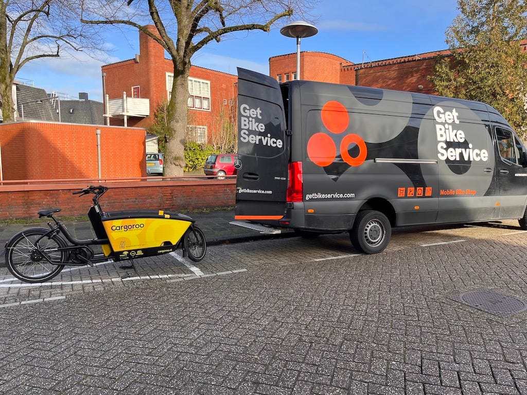 A Cargoroo branded cargobike parked behind a large van with its rear door open. The van is branded "Get Bike Service" who are a mobile bike repair company