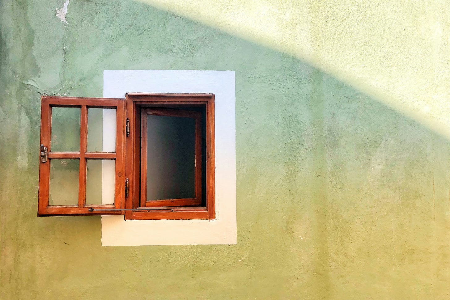 Muro verde chiaro con il sole che illumina obliquamente la parte in alto a destra. In mezzo, leggermente decentrata a sinistra, c'è una finestra di legno aperta, circondata da un rettangolo di pittura bianca lungo il contorno. 