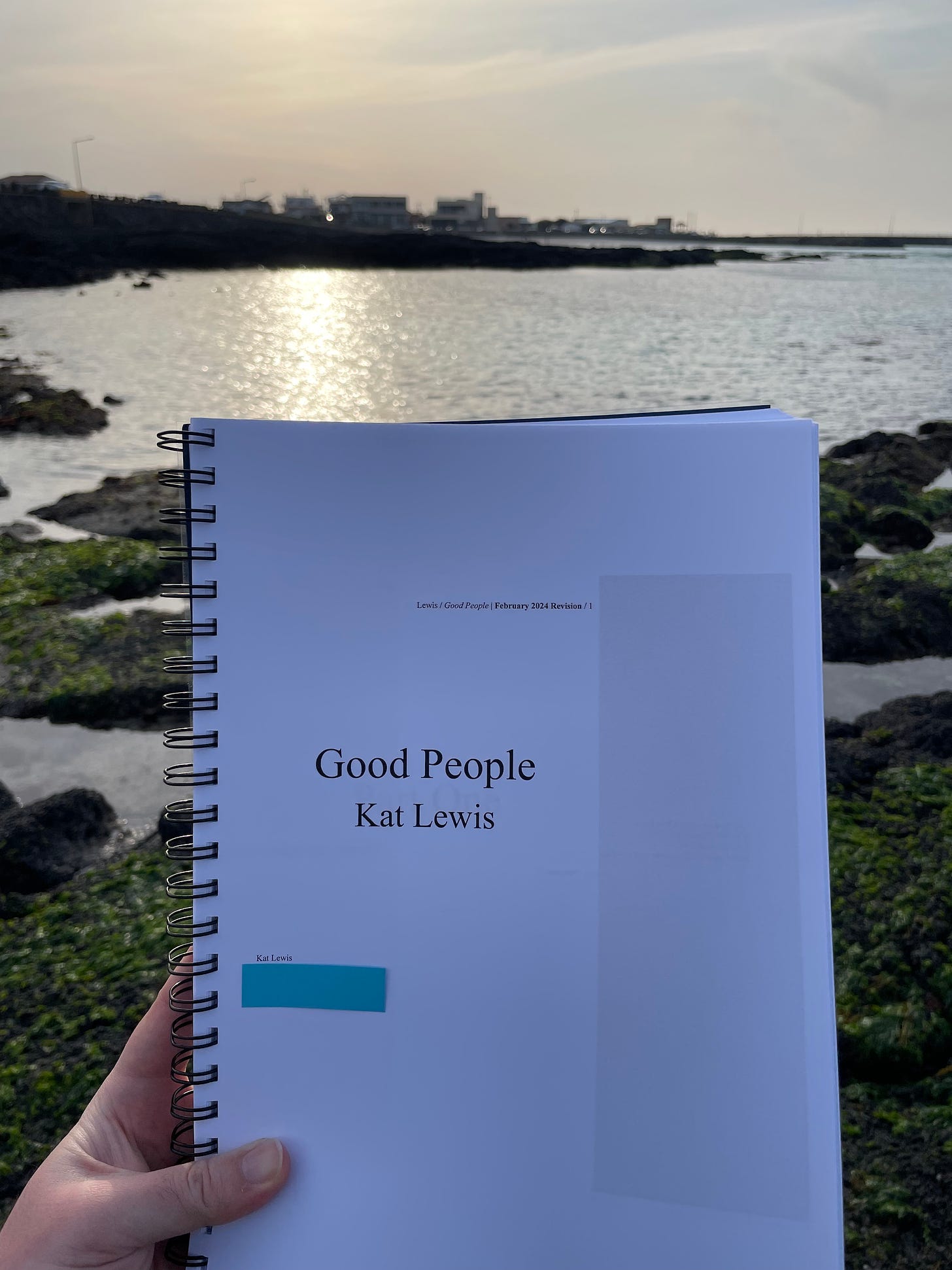 A photo of Kat's printed manuscript. The title page says Good People by Kat Lewis February 2024 Revision. A beach scattered with volcanic rock and tide pools is out of focus in the background.