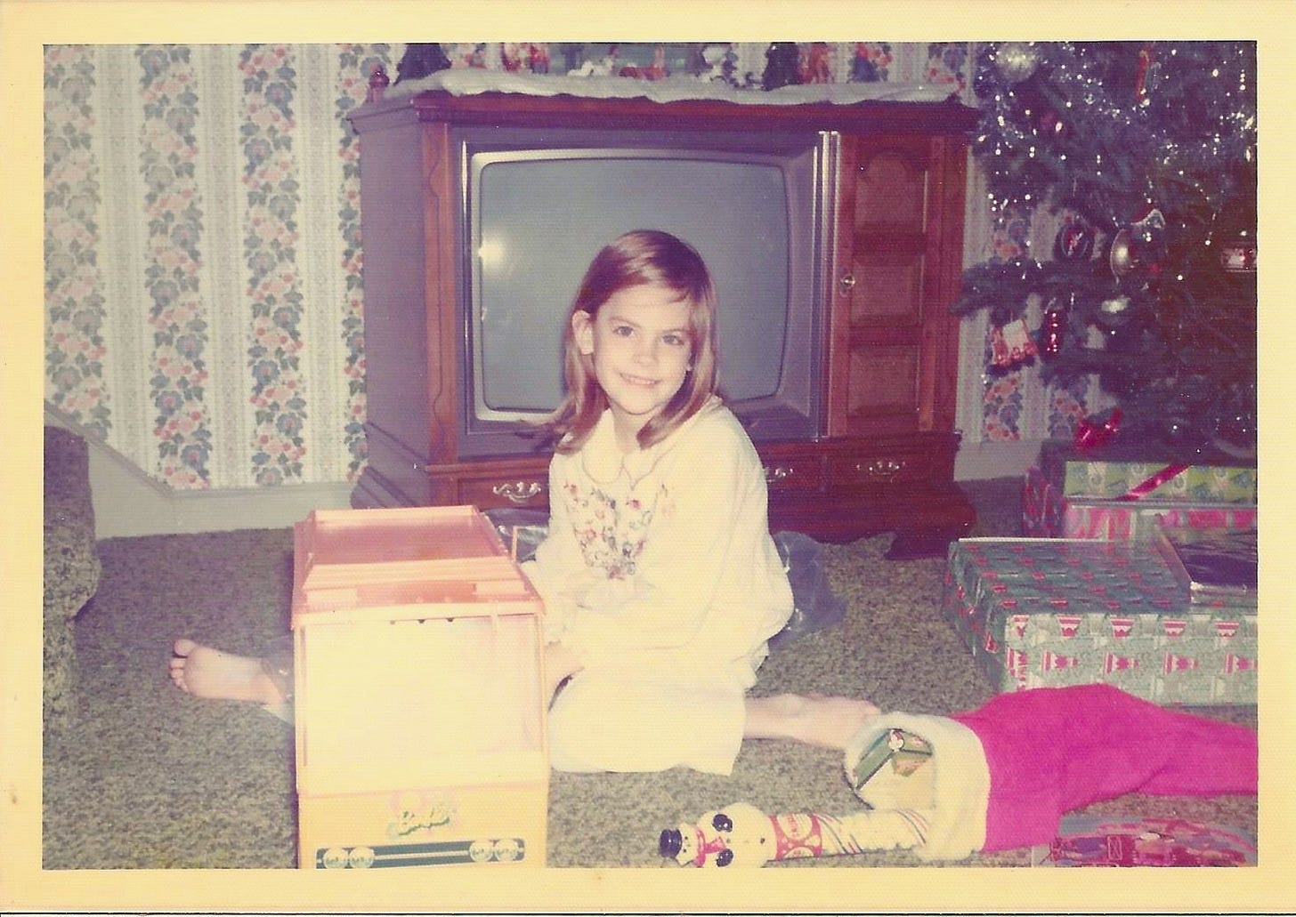 Little girl sitting beside a Christmas tree and all of her gifts, looking delighted that she's received a Barbie camper