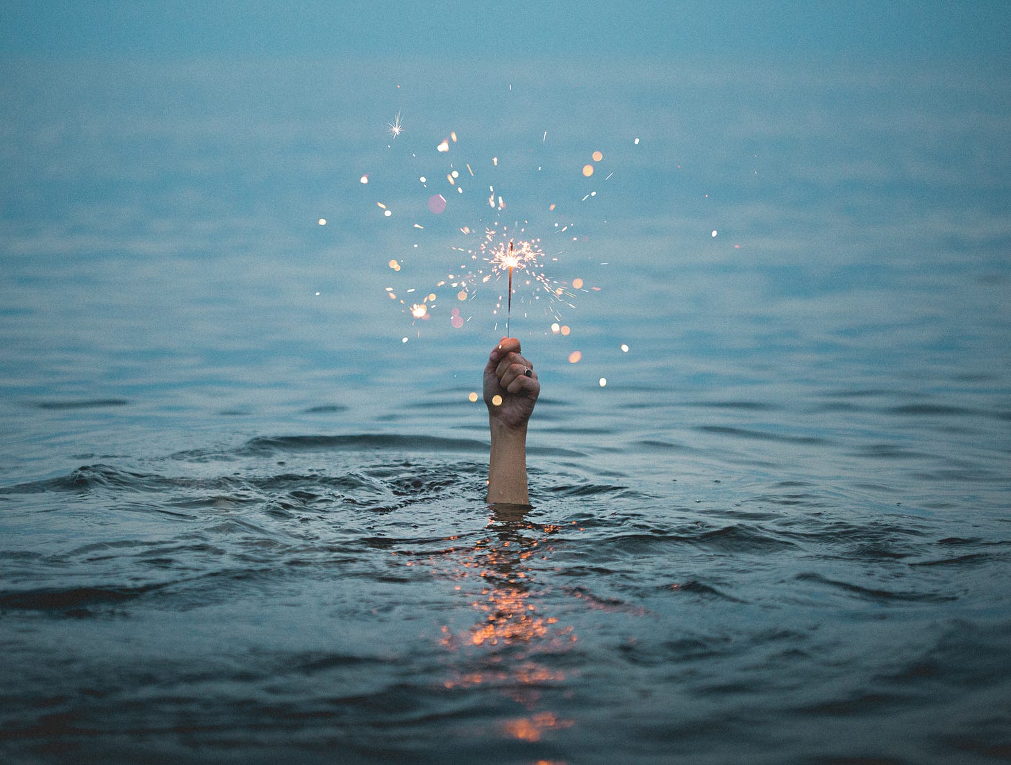 Mano che spunta dalla superficie del mare reggendo una candelina che fa scintille, tipo fuoco d'artificio.