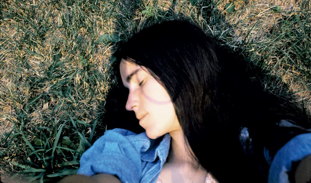 A young woman is lying on the grass, face in profile, dark hair flowing over her shoulders, eyes closed.