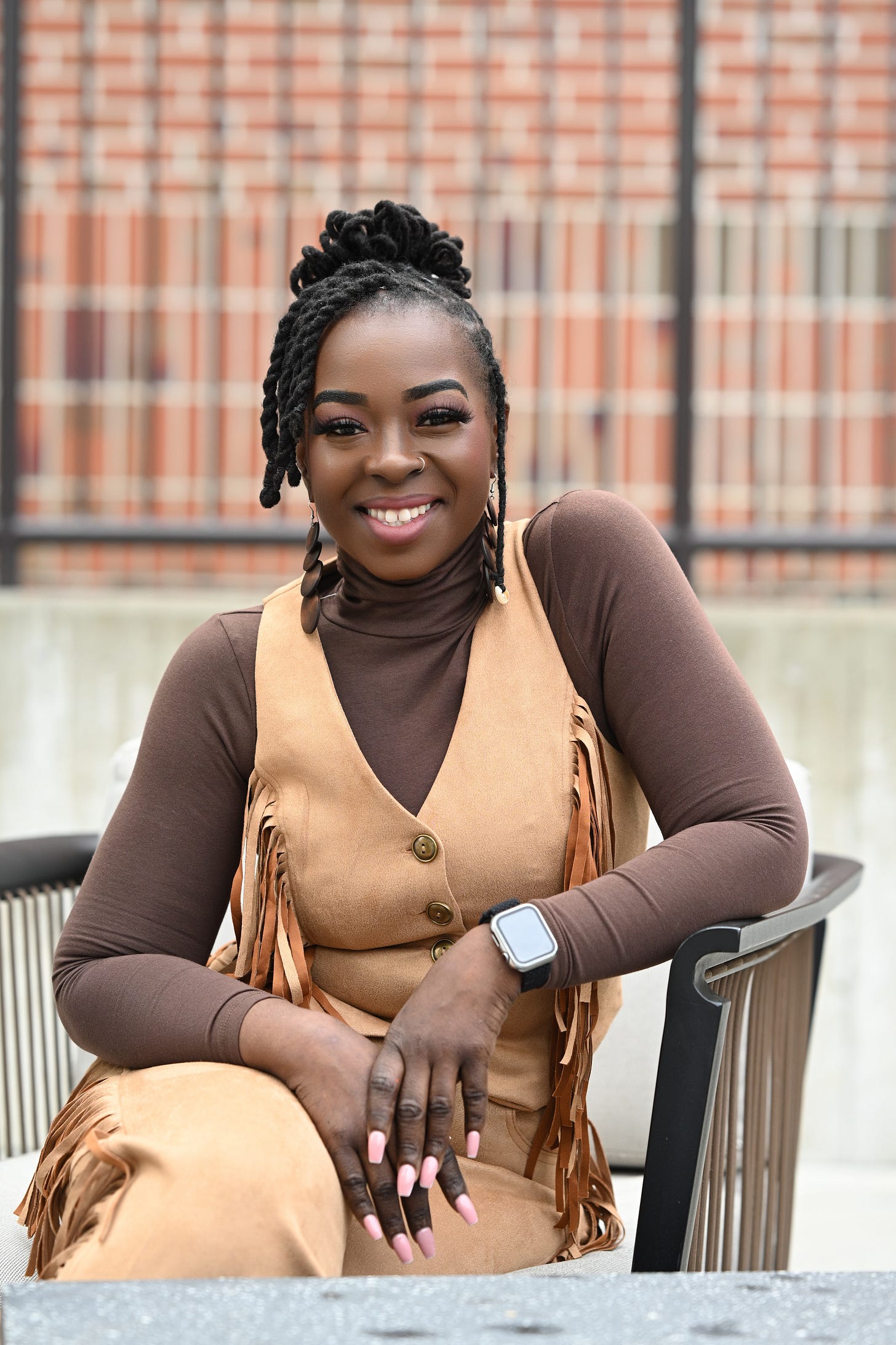 Monique McFadden, a chef, is seated at an outdoor table, smiling warmly. She is wearing a brown turtleneck under a tan suede vest with fringe details, paired with matching pants. Her hair is styled in elegant twisted locks, and she accessorizes with statement earrings and a smartwatch. The background features a blurred brick and metal fence. Photo by Nakeshia Shannon.