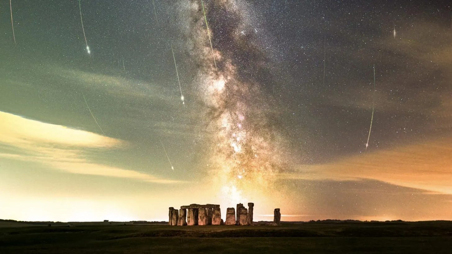 White fireballs streak over a darkened sky above the Stonehenge monument. The Milky Way ripples down through the centre of the sky. 