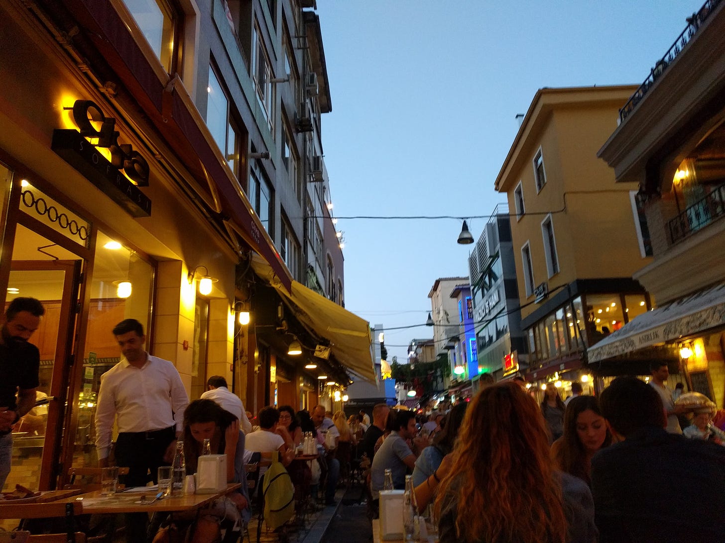 Diners sit at tables on the street outside a restaurant, under a darkening evening sky