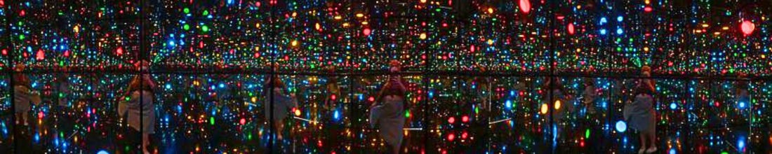 Kait inside Yayoi Kusama’s immersive installation, Infinity Mirrored Room—Illusion Inside the Heart (2020), at the New York Botanical Gardens in 2021. Dark mirrored room full of colorful dots of light. Multiple Kaits reflected.
