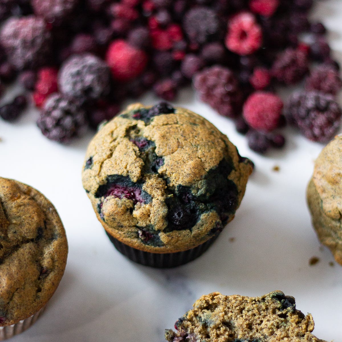 A berry muffin on a counter with a mound of frozen berries behind it.