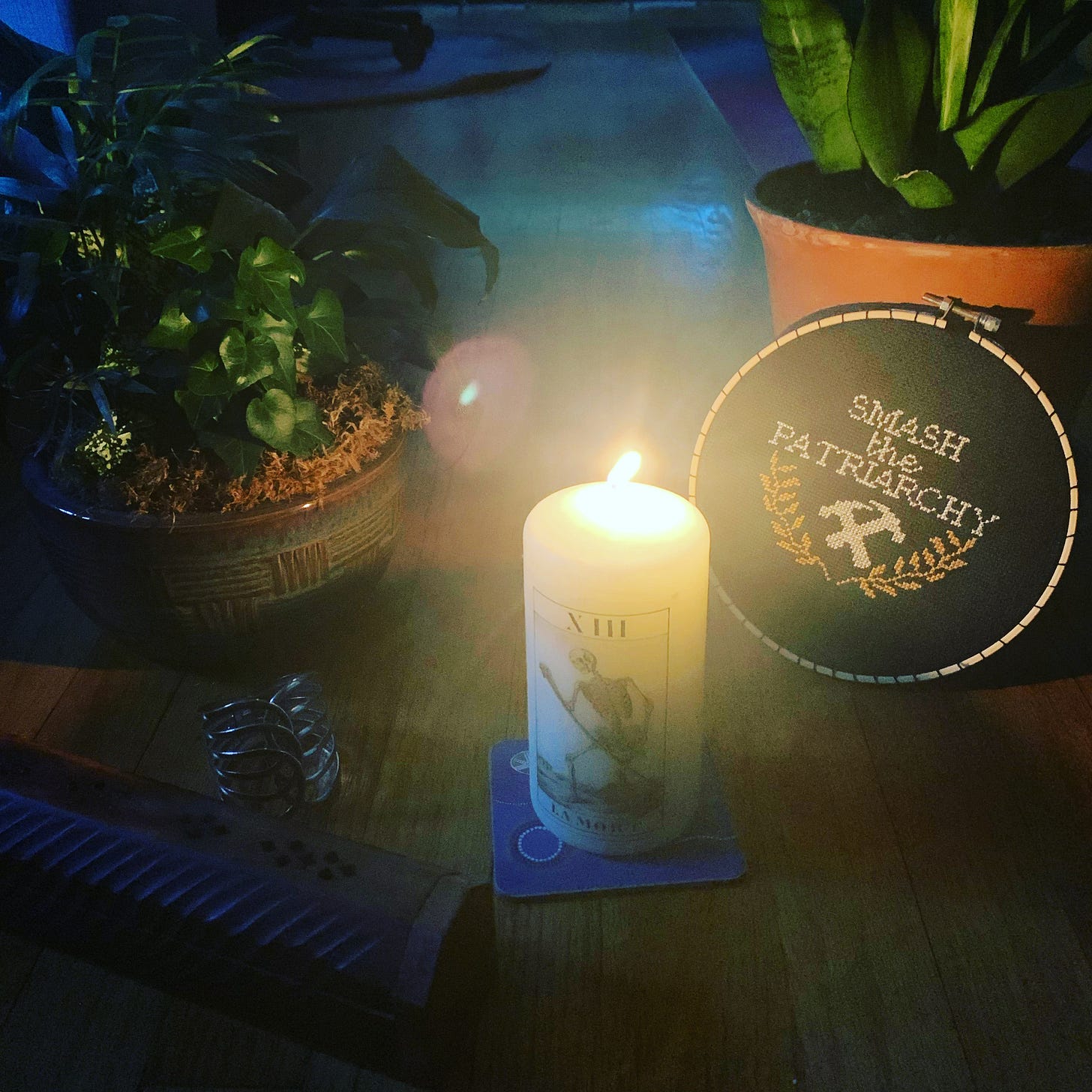 Two potted plants behind a lit white pillar candle. A cross stitch hoop to the right says smash the patriarchy in white letters.