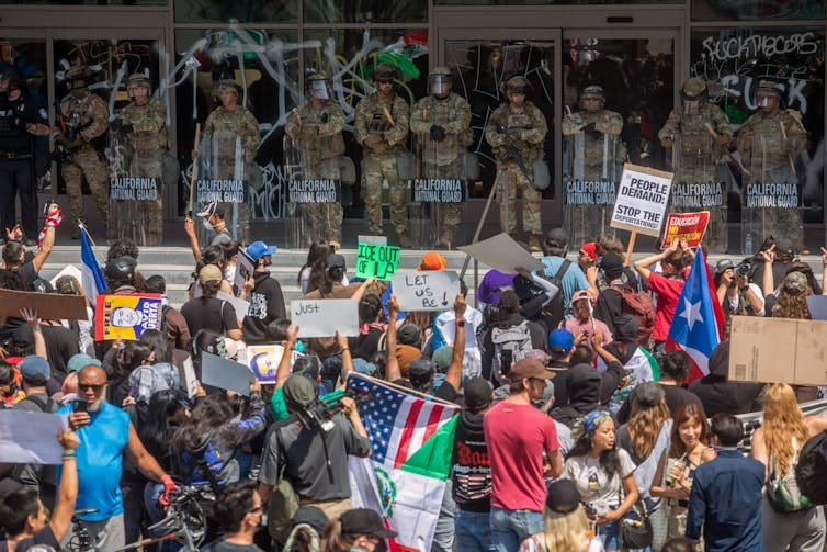 A large crowd of people hold flags and face a row of people dressed in camouflage, standing above them on steps. A large crowd of people hold flags and face a row of people dressed in camouflage, standing above them on steps.