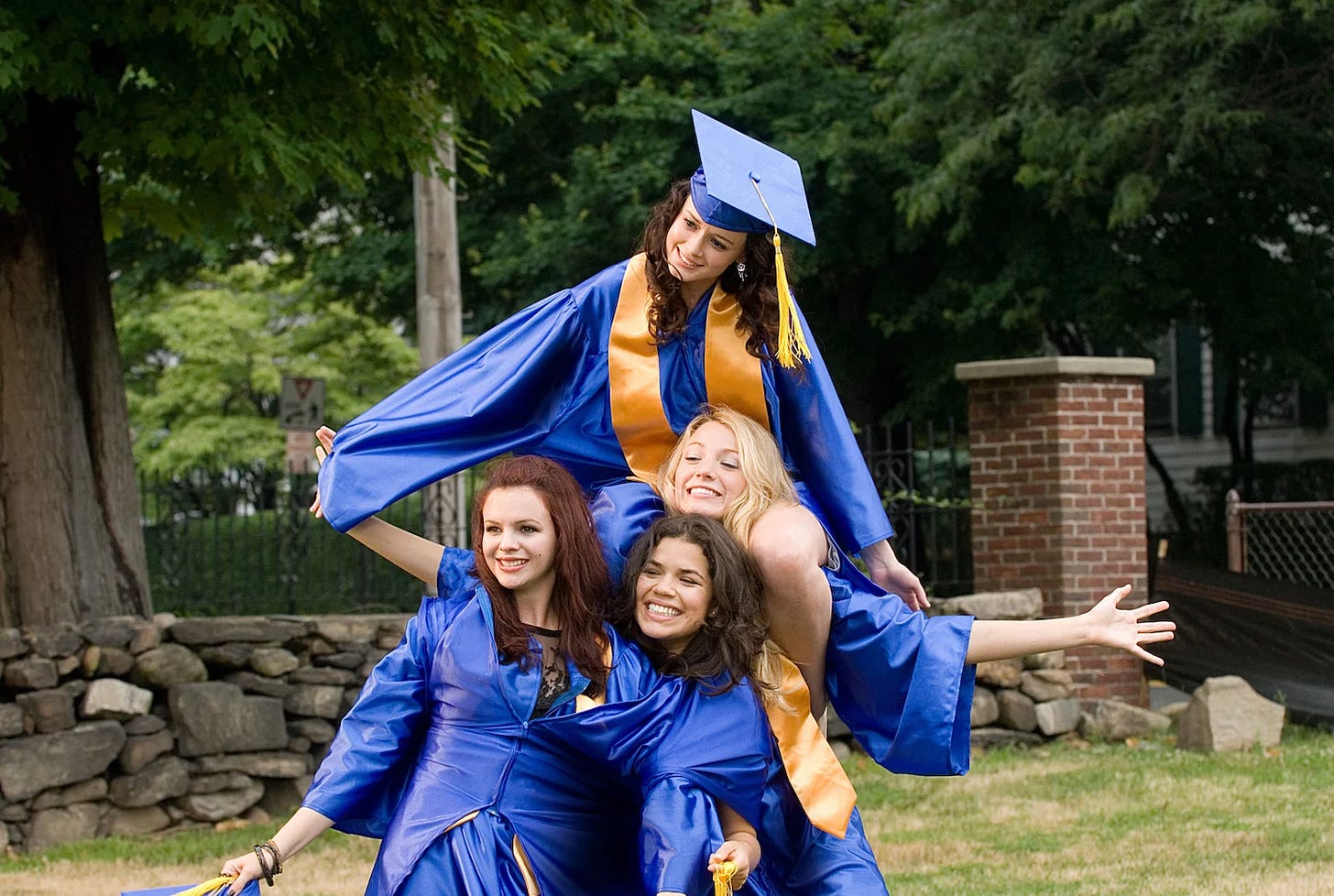 A still from the movie "The Sisterhood of the Traveling Pants 2." The four main characters, Tibby (Amber Tamblyn), Carmen (America Ferrera), Lena (Alexis Bledel), and Bridget (Blake Lively) are all in blue graduation robes posing for a photo. Tibby and Carmen have each put one arm through the sleeve of another robe so that they are both zipped inside a robe made for one person. Behind them, Lena sits on Bridget's shoulders. They all smile.