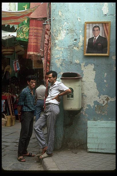 Local men chatting by Pres. Ben Ali's portrait-graced wall in fore of colorful, ware-lined souk .