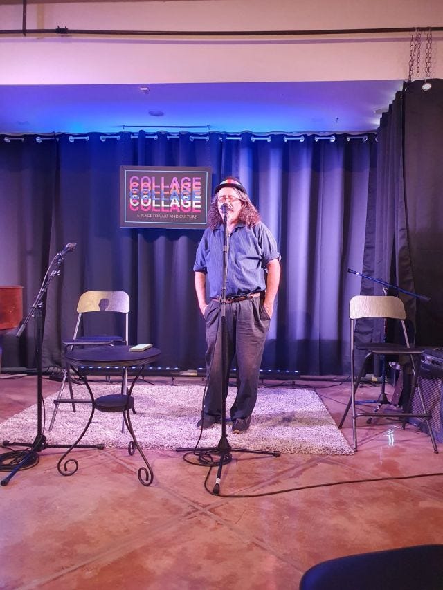 a photo of my dad giving a standup/philosophy lecture. A tanned white guy with long dark hair and dark clothing, glasses and a rainbow hatband, standing in front of a purple-lit curtain and a sign reading "collage: a place for art and culture"