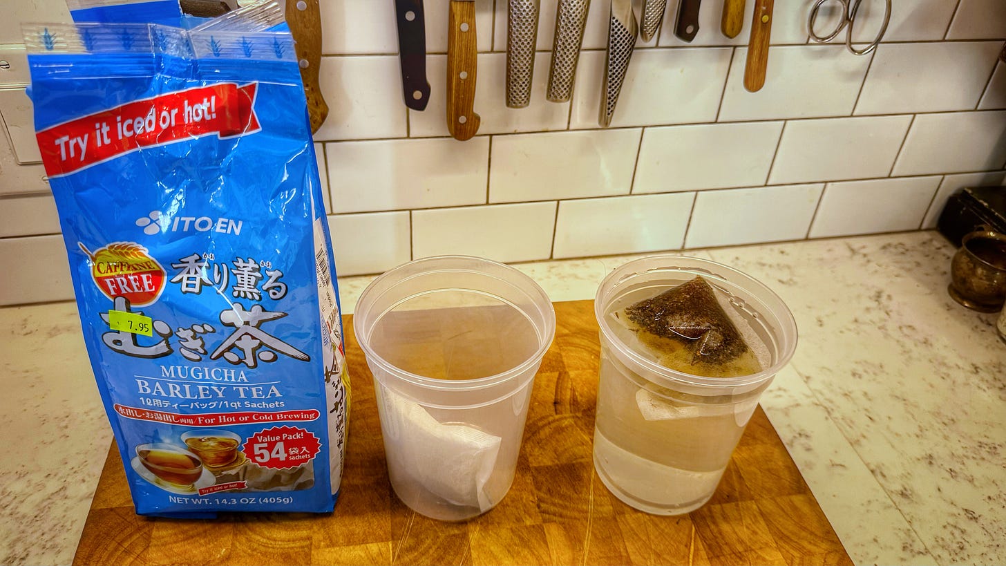 A package of 54 mugicha barley tea bags beside two plastic quart containers. One holds only a dry tea bag. The other is filled with water with a tea bag floating in it. All of this is also on the cutting board on the counter. 