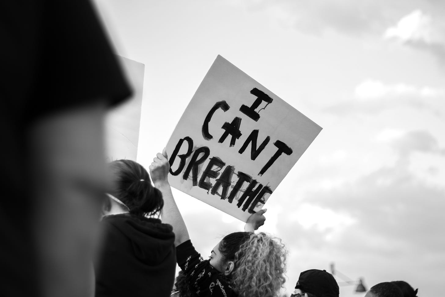 A black-and-white photo of a young woman holding up a protest sign against a cloudy sky, surrounded by a crown of people. The sign, written with black marker on white cardboard, reads "I CAN'T BREATHE"
