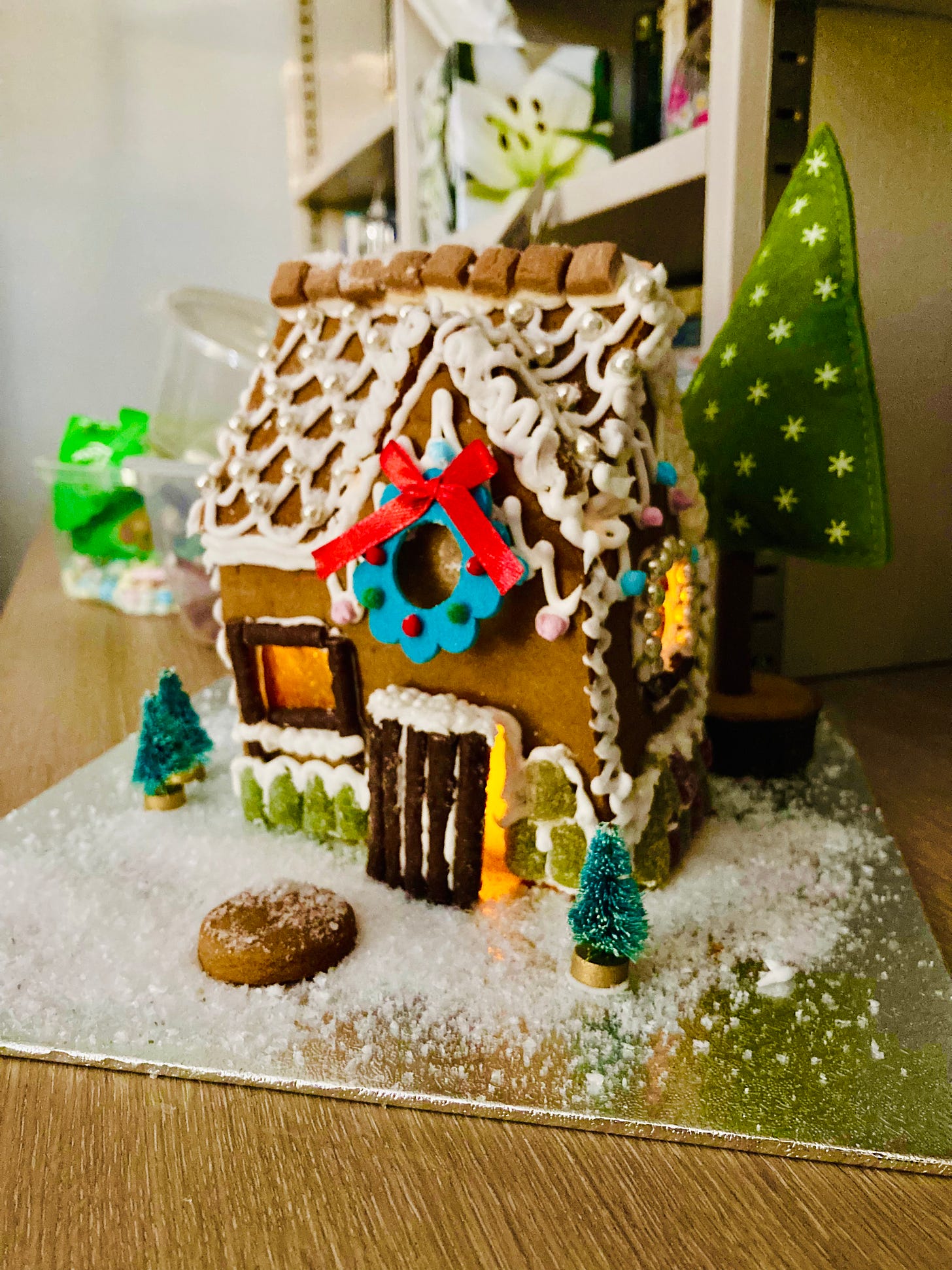 photograph of a small gingerbread cottage, with loopy white icing, pink and blue millions, green midget gem decorations, and chocolate matchstick window panes. Organge light shines through the windows and half-open door and the house is surrounded by fake white snow, three tiny christmas trees and a giant felt christmas tree. A felt wreath hangs on the front above the door.