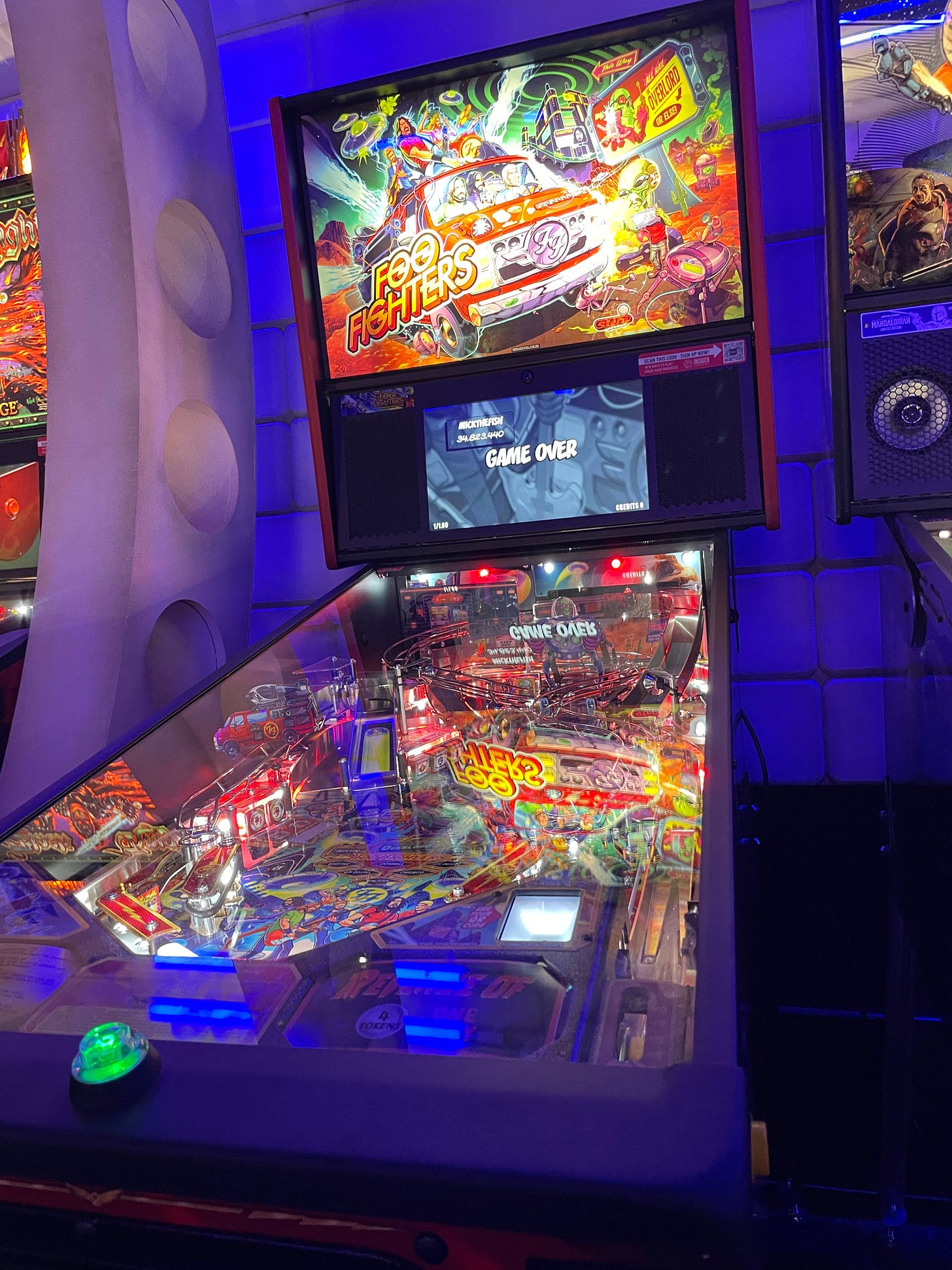 A photo of a very colorful Foo Fighters pinball machine in a small room designed to look like the interior of a spaceship with purple lighting.