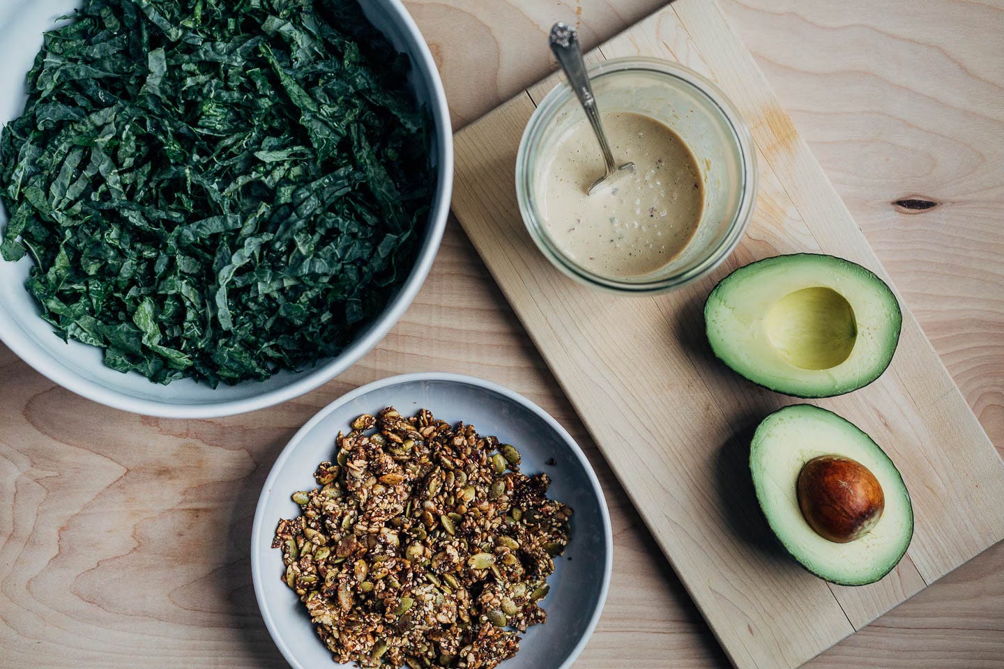 Ingredients laid out: kale in a bowl, dressing and an avocado, and a bowl of seed clusters