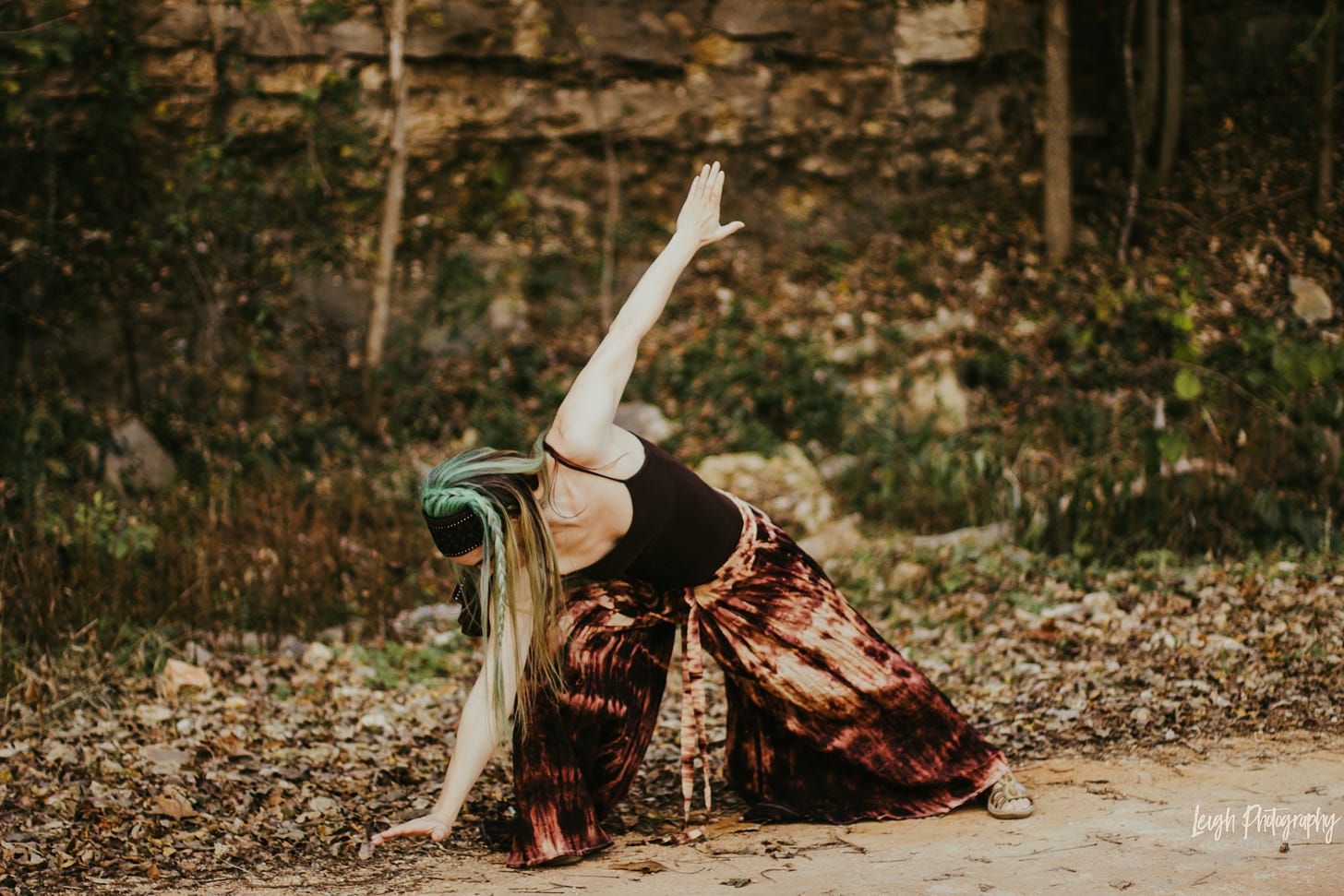 The green-haired author in earth tone costume dancing on dirt before a towering rock wall and green shrubs.