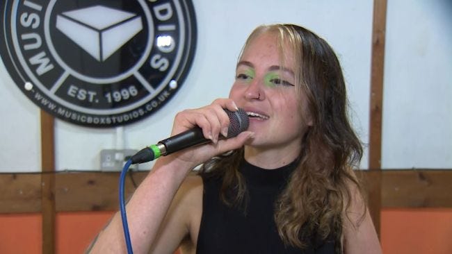 A femme performer is singing into a mic, wearing a black sleeveless shirt and striking green eye makeup, with long wavy brown hair.