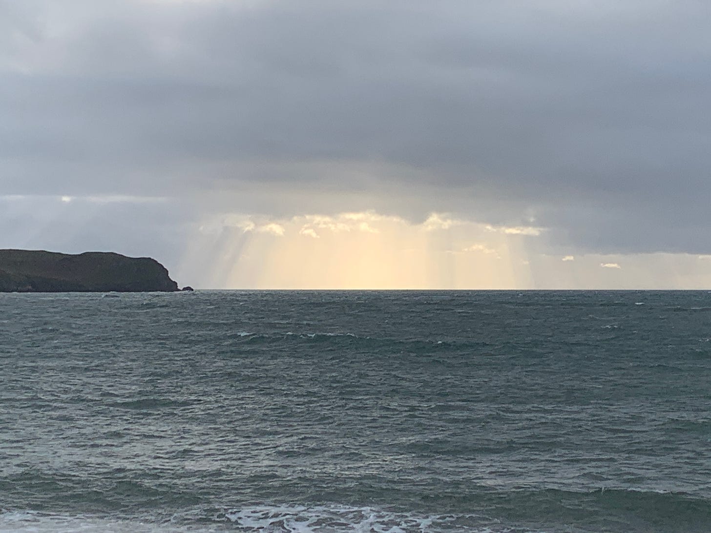 Ocean, with grey skies and a breakthrough column of bright sunlight through the clouds. A headland is visible to the left