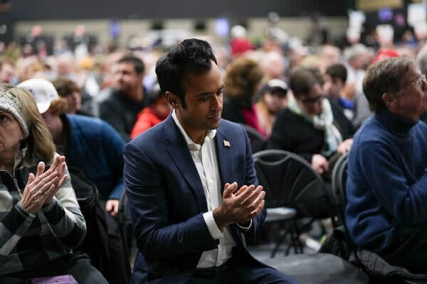 Vivek Ramaswamy sits and claps at a caucus site, surrounded by a crowd of people also in seats.