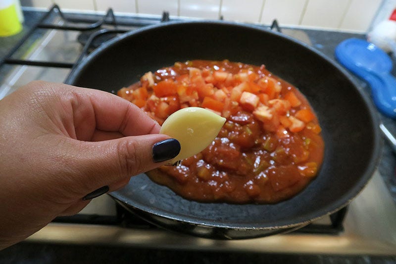 A hand holds a piece of garlic in front of a pan of sauce.