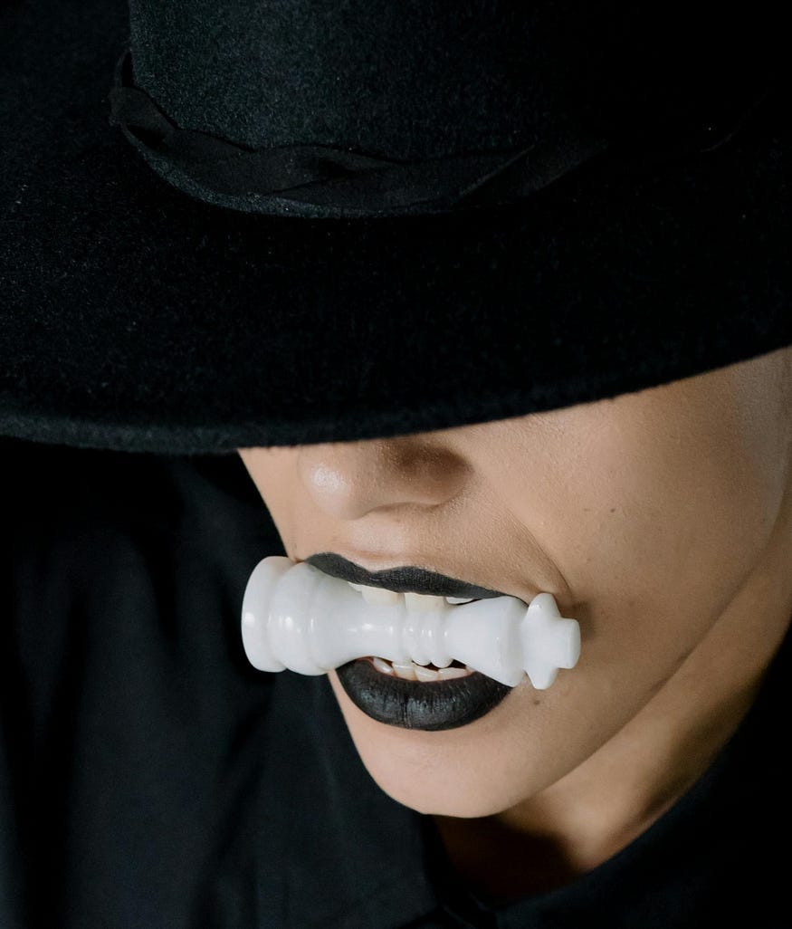 A woman wearing dark-colored lipstick bites down on a white chess piece. The upper part of her face is covered by a black hat. A woman wearing dark-colored lipstick bites down on a white chess piece. The upper part of her face is covered by a black hat.