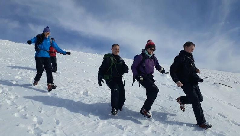 Four people run down an extensive bank of snow with big grins. There is a fifth person partially hidden behind the left-most person. Everyone is full of dynamism and fun. It is a bright day, with a blue-and-white sky in the background. 