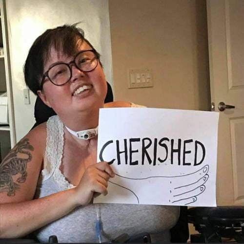 A photo of Stacey Park Milbern, a mixed race Korean and white queer person, holding a paper sign that reads “Cherished.”