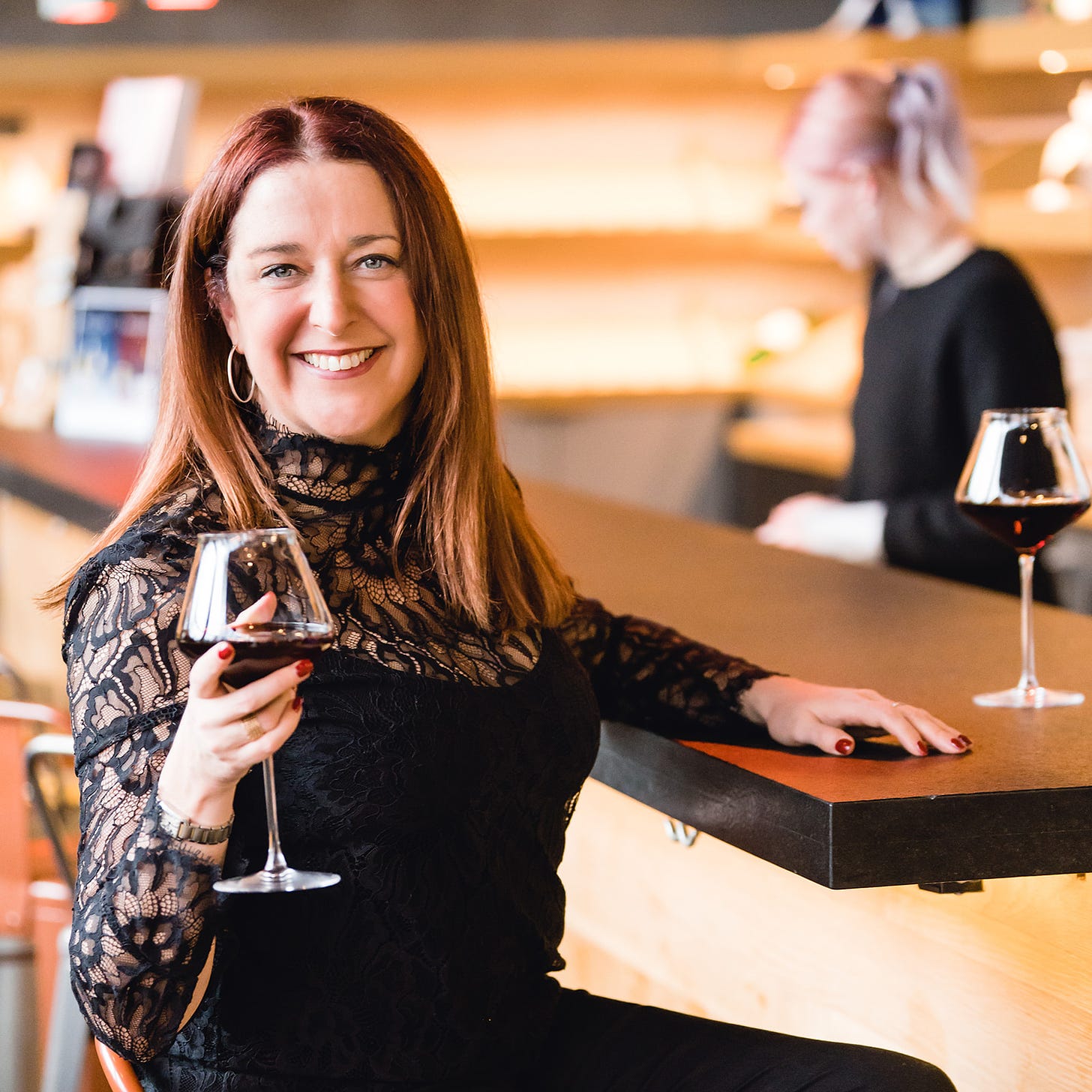 A woman dressed in black sitting at a bar holding up a glass of red wine. 