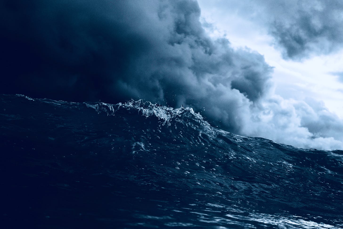 blue-tinted black-and-white photograph of a cresting wave against a backdrop of ominous stormclouds