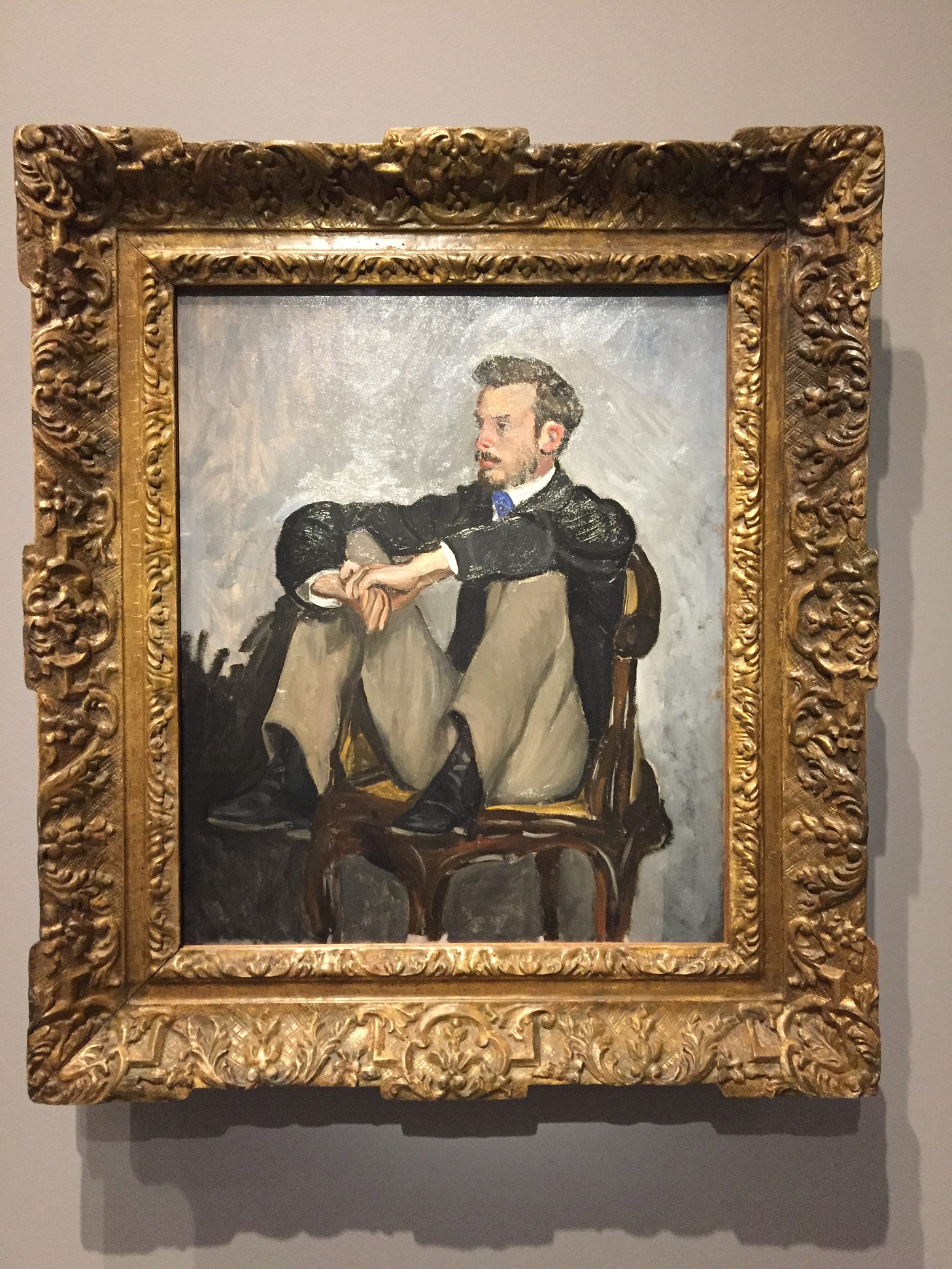 Frédéric Bazille’s portrait of Pierre-Auguste Renoir. He is sitting on a chair with his legs drawn.