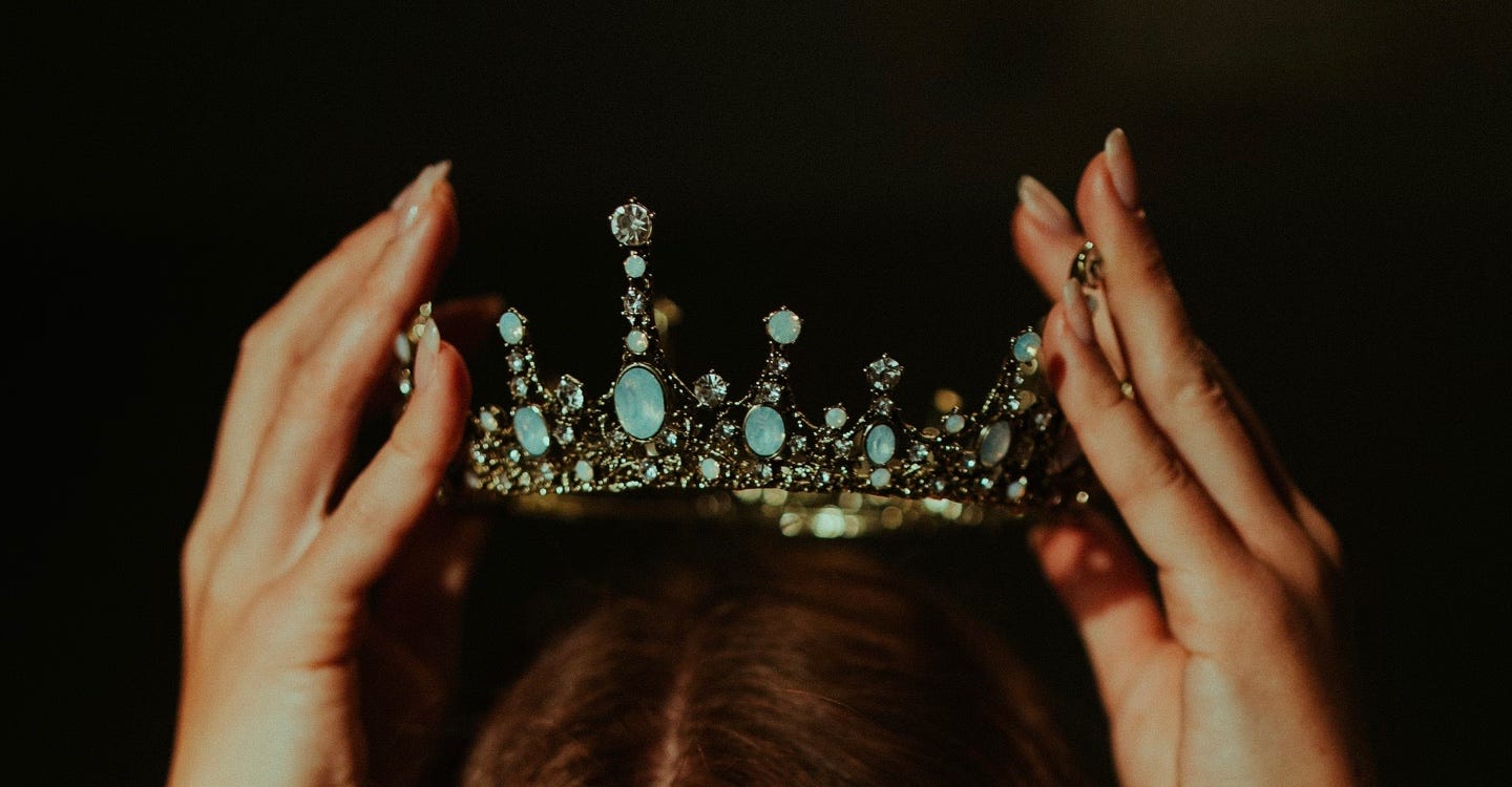 Two female hands are pictured in close up holding a silver crown, set with blue stones, above a head of brown hair.
