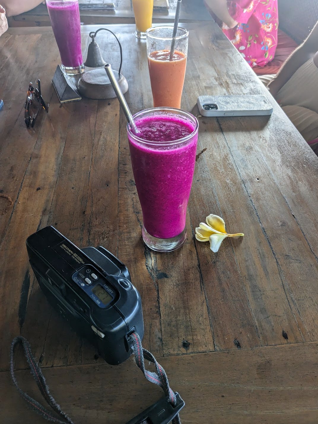 a bright purple / fuscia drink lies next to a white and yellow flower and black point and shoot camera on a wooden table