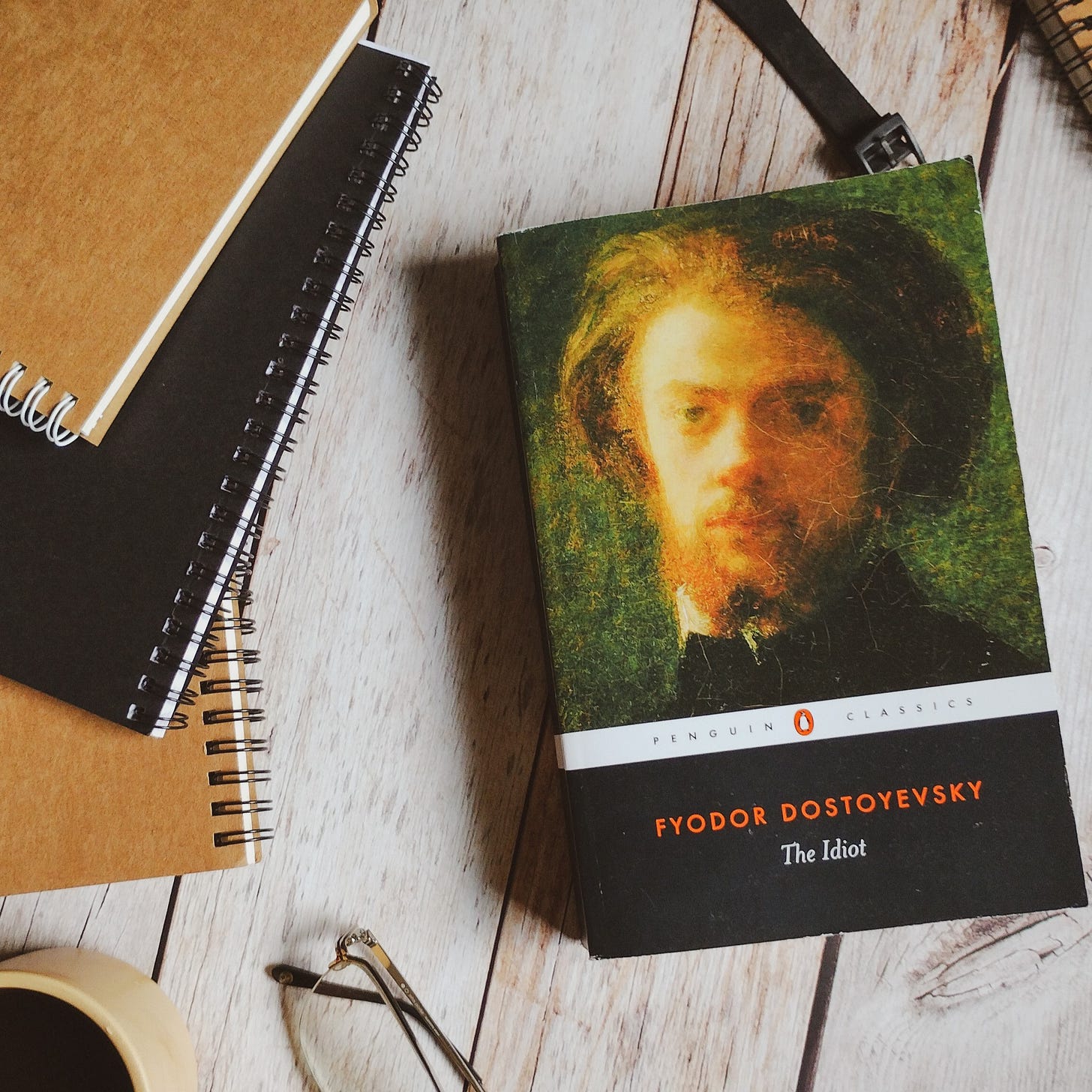 The Idiot by Fyodor Dostoevsky, on a wooden tabletop, alongside some notebooks 